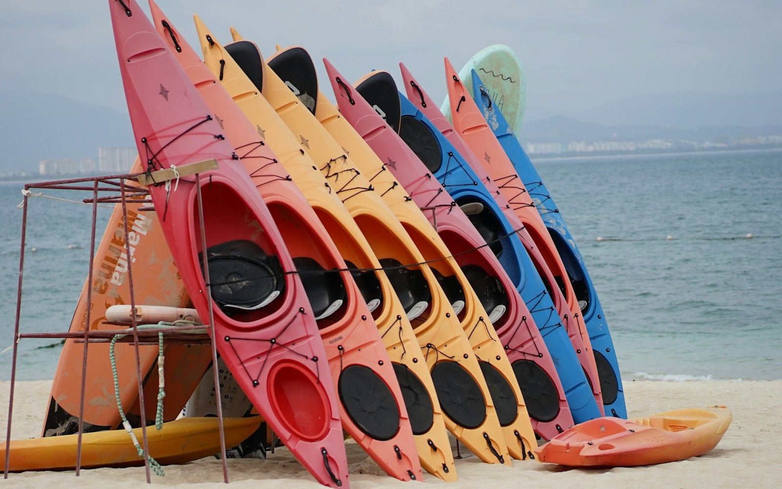 Kayaks sit-on-top de plusieurs couleurs rangés debout sur un râtelier, au bord d'une plage