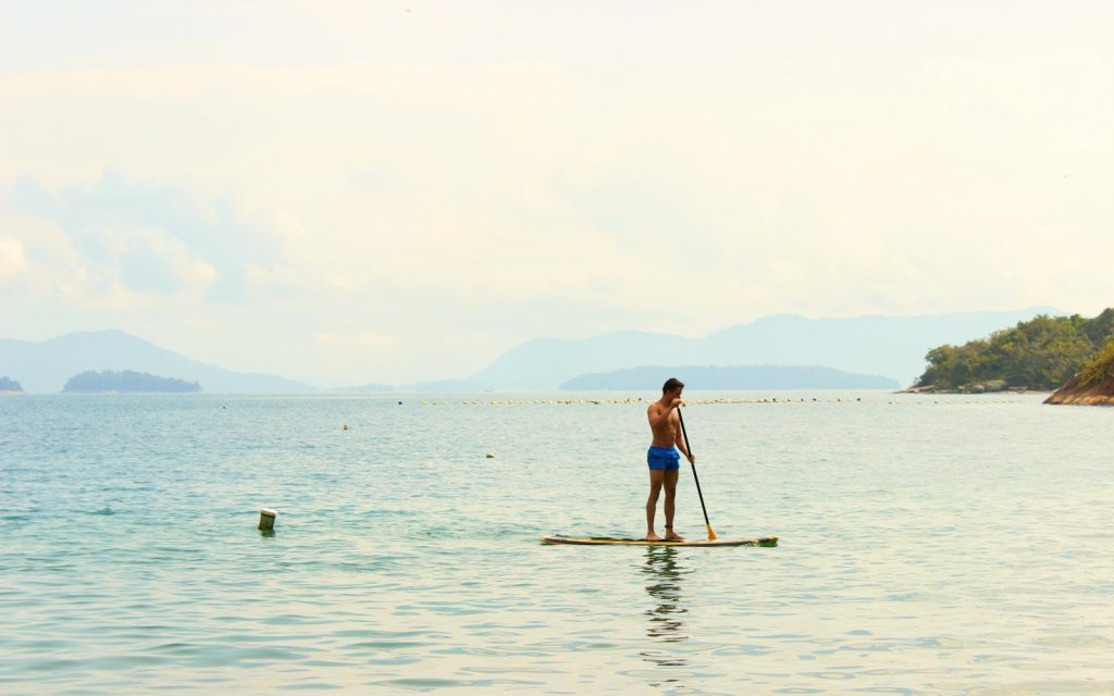 Un pagayeur debout sur sa planche près du rivage, sur une mer calme, avec des collines à l'horizon