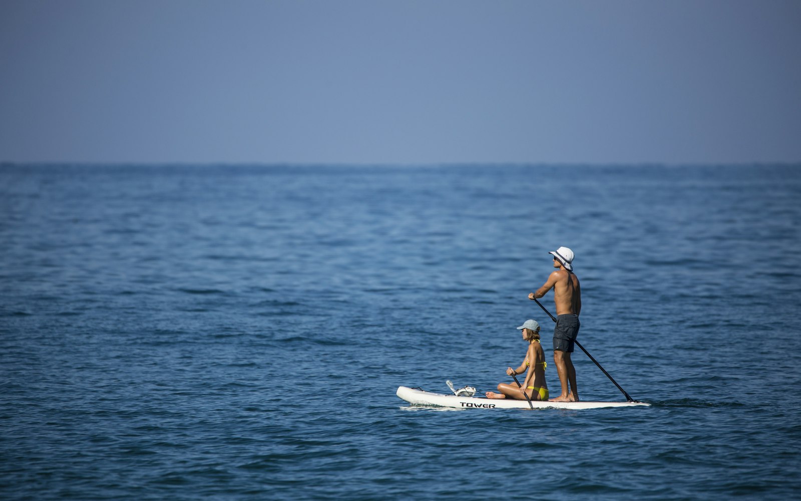 Un adulte debout et un enfant assis sur un stand up paddle, sur une mer bleue et calme