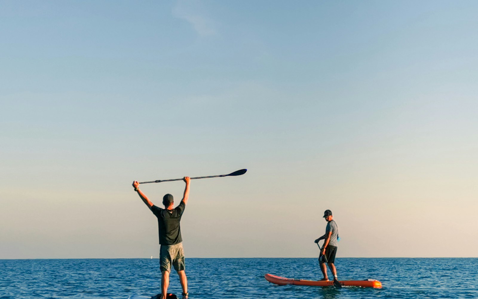 Deux pratiquants de stand up paddle sur une mer calme, l'un levant sa pagaie, sous un ciel dégagé
